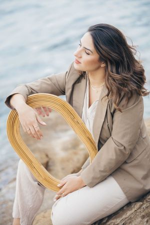 Woman in beige blazer poses with circular golden frame against coastal backdrop.