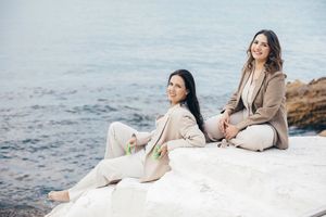 A couple in white outfits sits together on coastal rocks by the ocean during a romantic photoshoot.