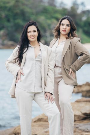 Two women in neutral-toned outfits pose together on rocky coastline during outdoor photoshoot.