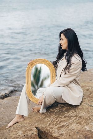 A woman in a beige outfit sits on coastal rocks holding a decorative gold mirror by the ocean.