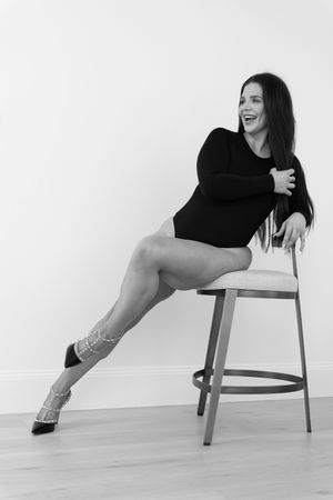 Artistic black and white photo of a dancer in black leotard posing joyfully on a stool against white wall.