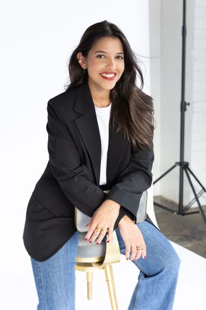 Professional studio portrait of someone in black blazer and jeans sitting on gold stool.