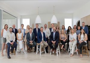 A professional business team poses together in a bright modern office space with white pendant lights and windows.