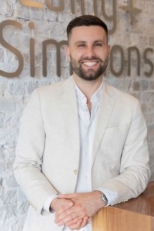 Professional wearing a white blazer and button-down shirt sits at wooden table against textured wall.