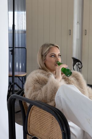Person in fur coat sits in cane chair while drinking from green bottle.