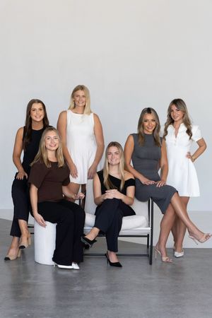 A group of women in black and white outfits pose casually together against a gray studio backdrop.