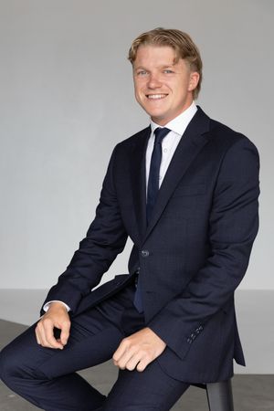 Professional headshot of someone wearing a dark navy suit and tie sitting casually against a light gray background.