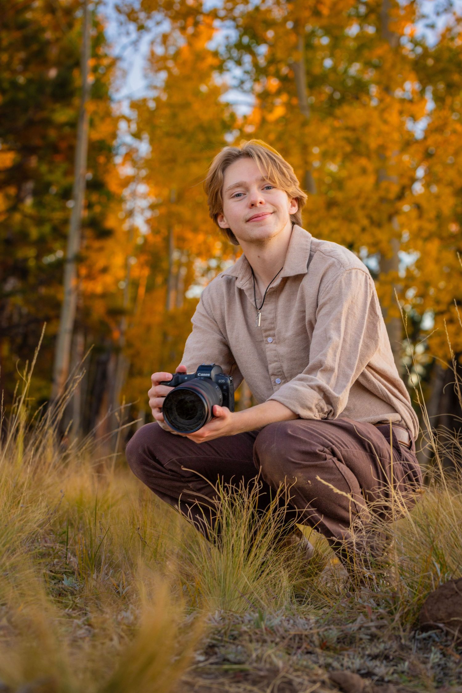 Chandler crouching in tall grass holding a Canon R5 camera surrounded by vibrant golden autumn aspen trees.