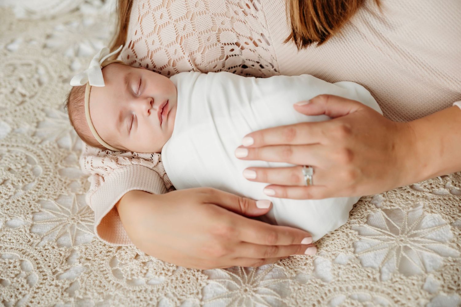 Tender hands gently cradle a sleeping newborn baby wrapped in soft white fabric against a textured beige background.  Middle TN Photographer