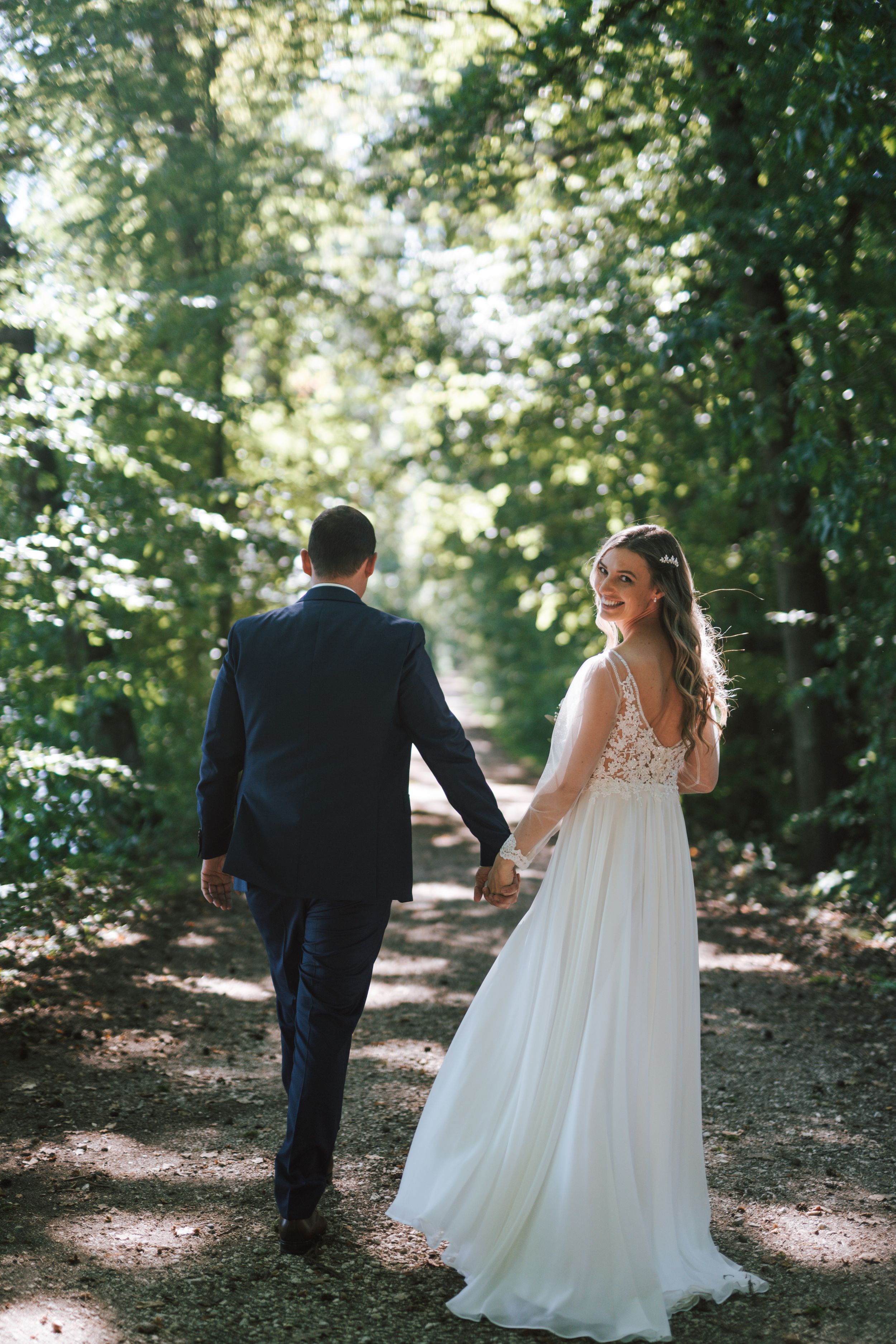 A bride in a flowing lace dress and groom walk along a tree-lined woodland trail.