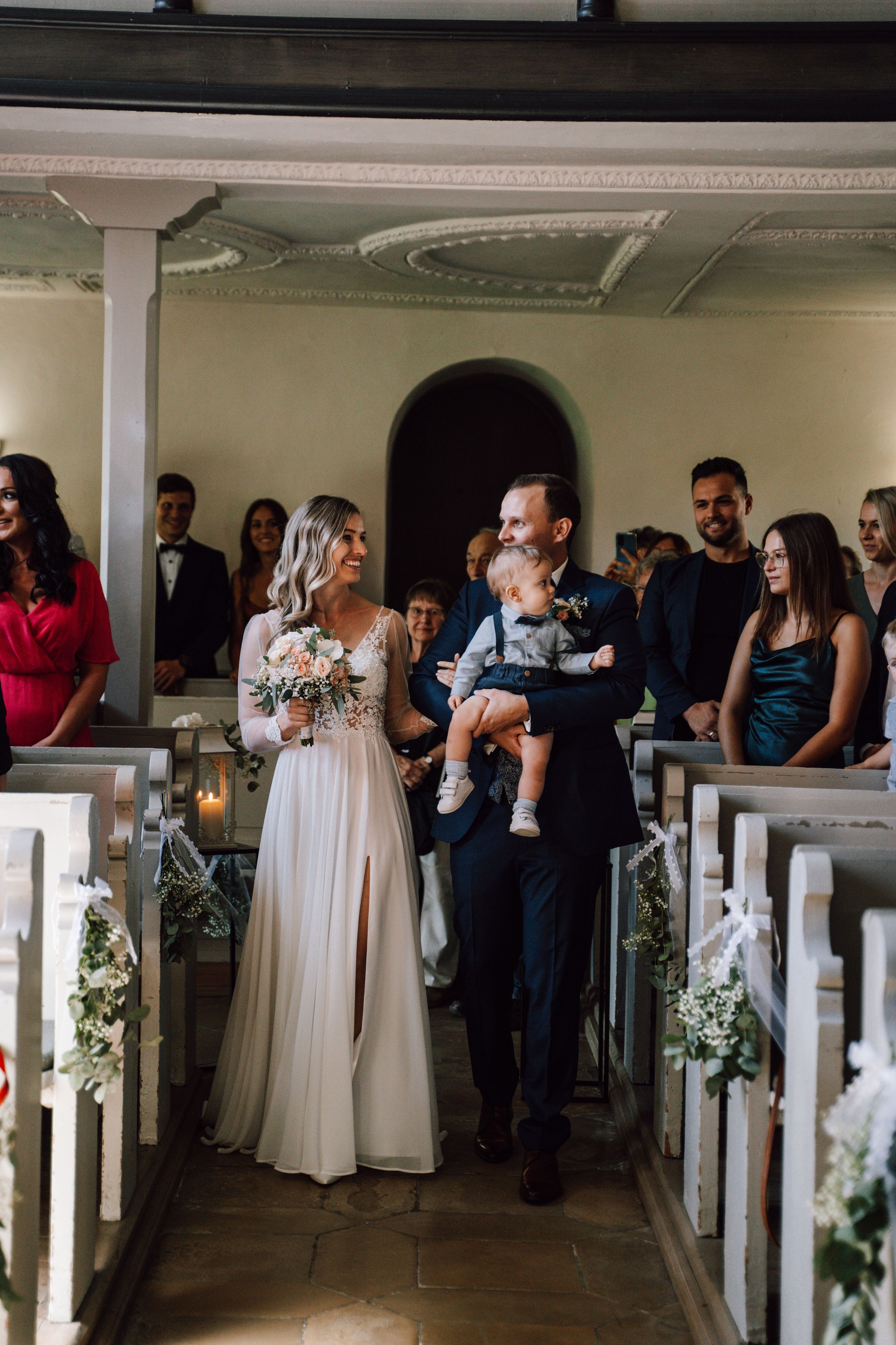 Wedding guests gather in elegant indoor venue with white railings and floral decorations.