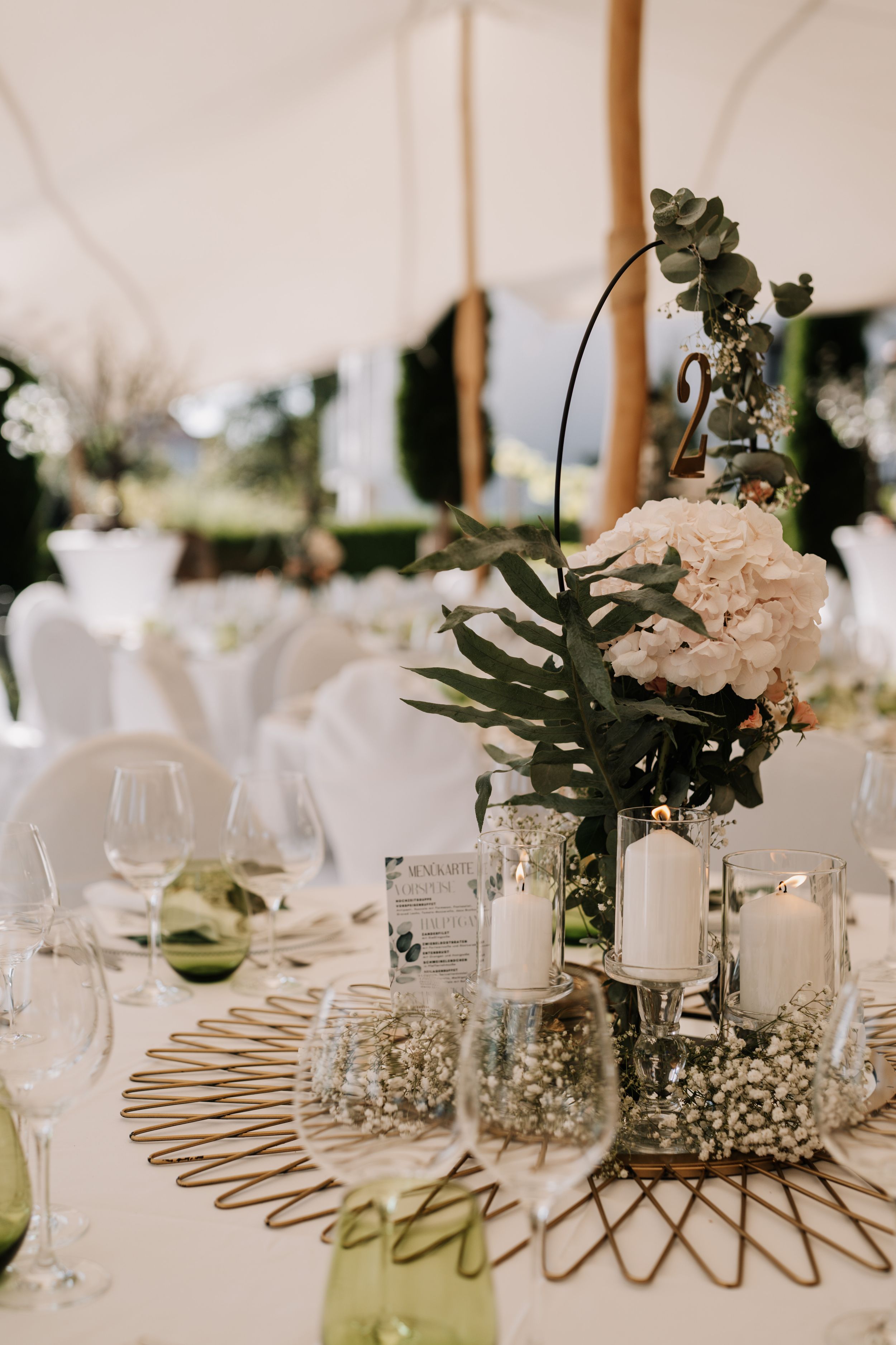 Wedding reception centerpiece featuring white candles and greenery on a metallic decorative base.