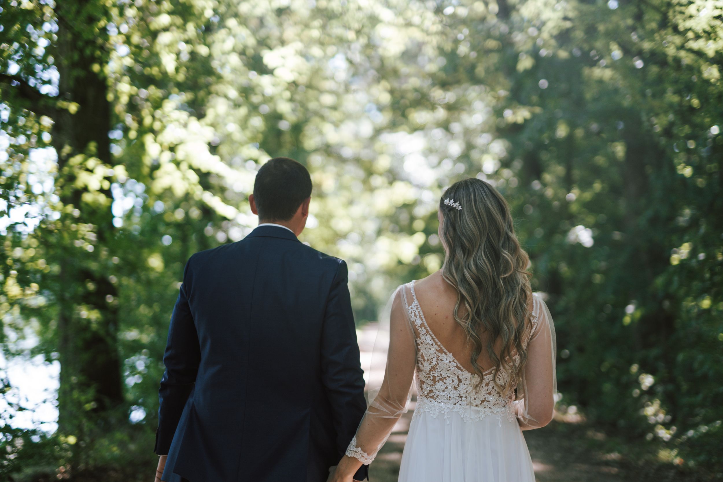 A couple in wedding attire walk hand in hand through a sunlit forest path.