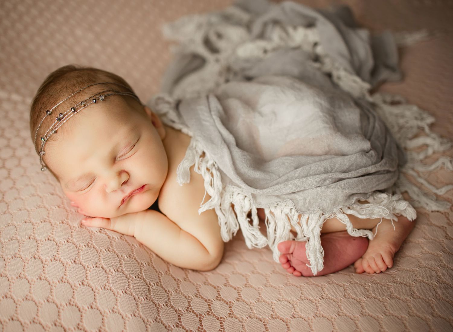 Newborn baby sleeping peacefully wrapped in gray textured blanket on pink surface.