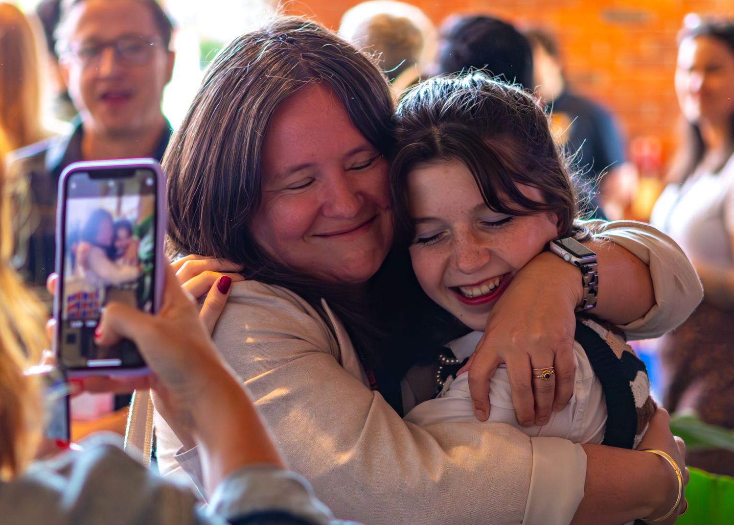 Two attendees share an emotional embrace while someone captures the moment on a phone.