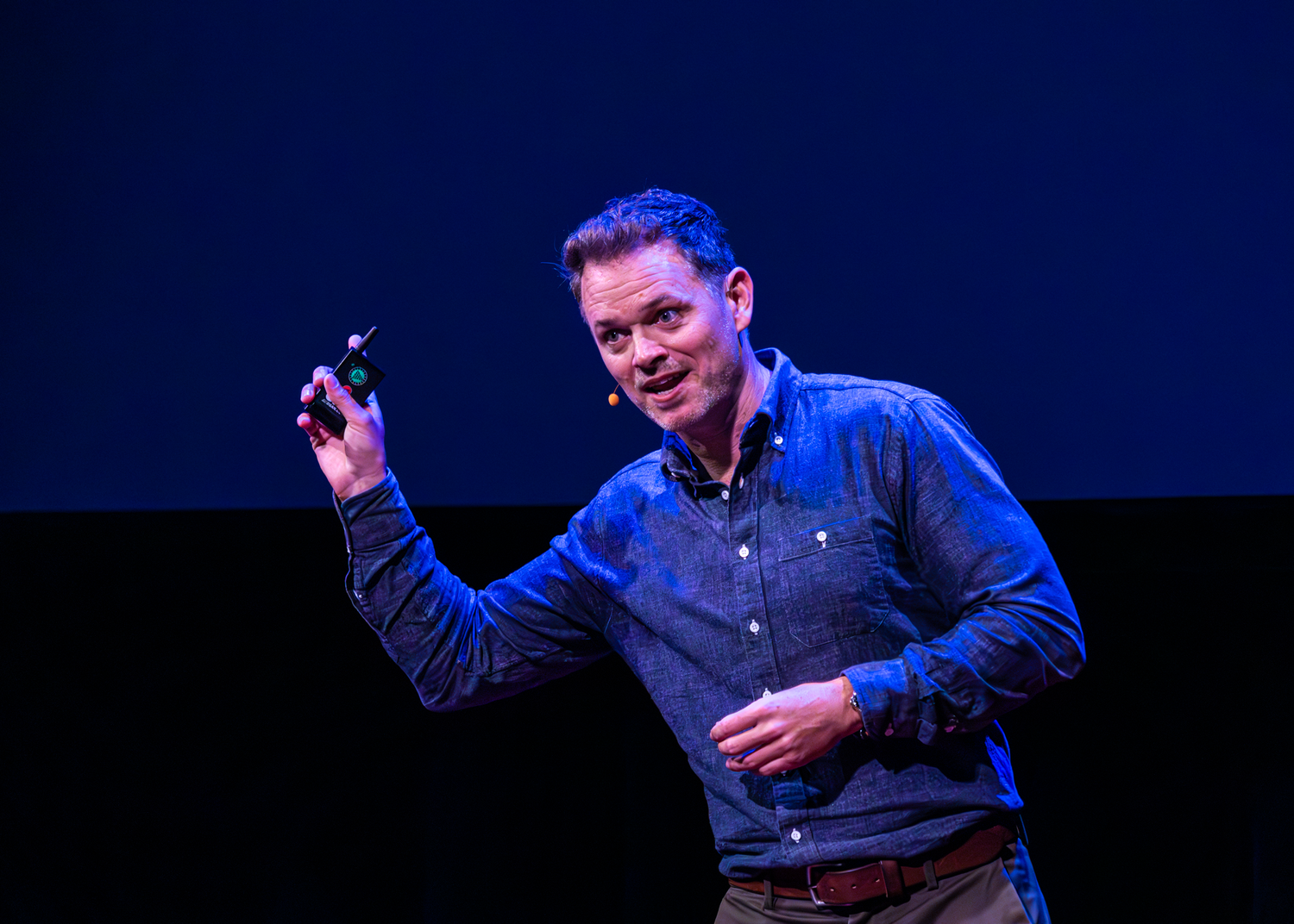 A speaker gestures enthusiastically while presenting on the TEDx stage with blue lighting.
