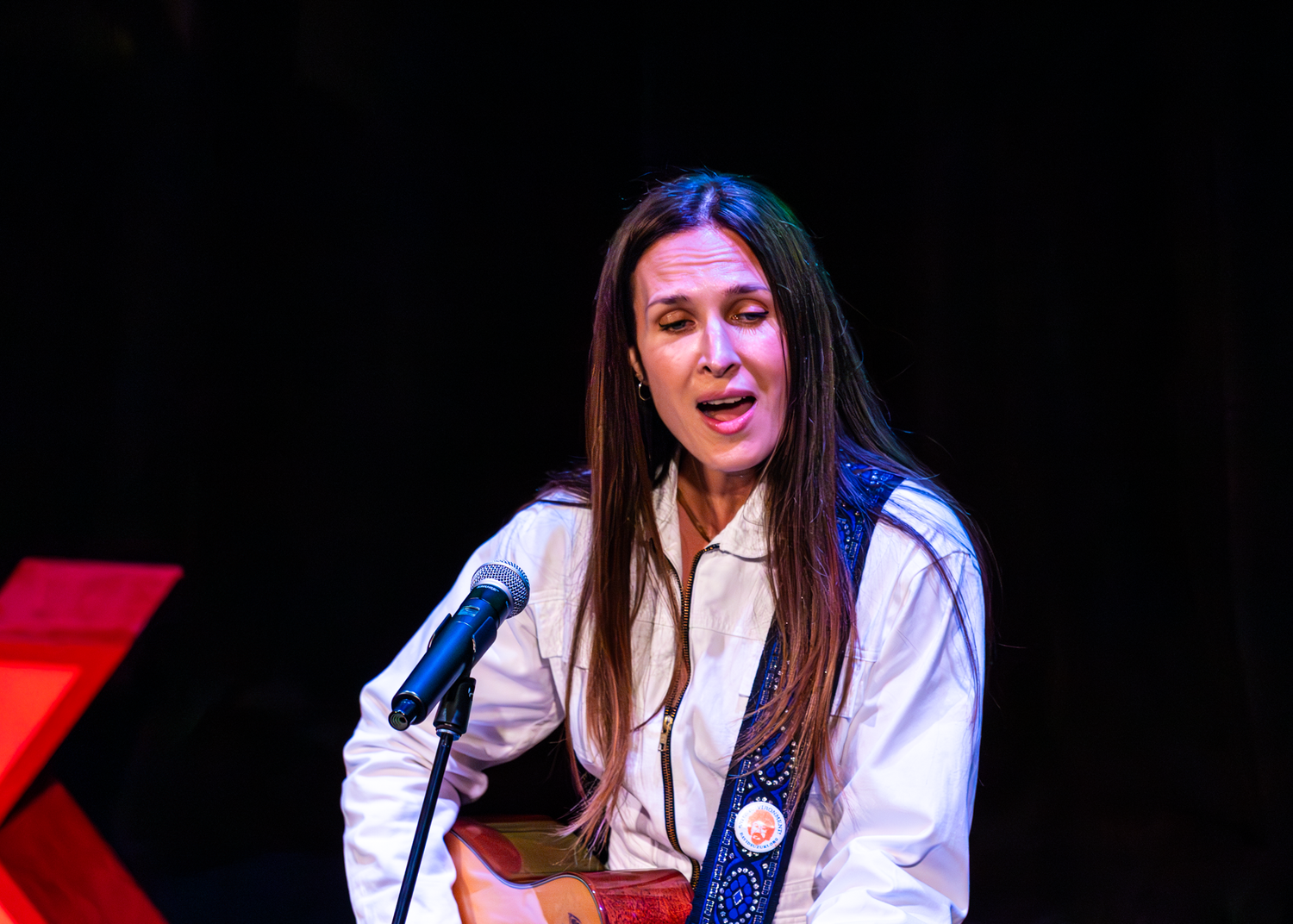 A performer sings on stage at TEDx Victoria while playing an acoustic guitar.