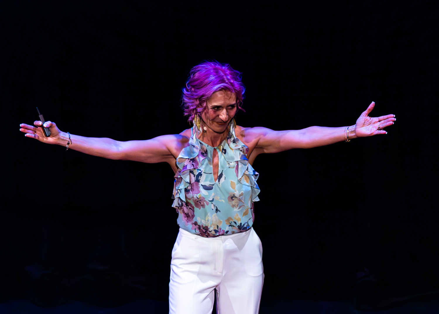 A performer in a floral top and white pants stands with arms outstretched on the TEDx stage.