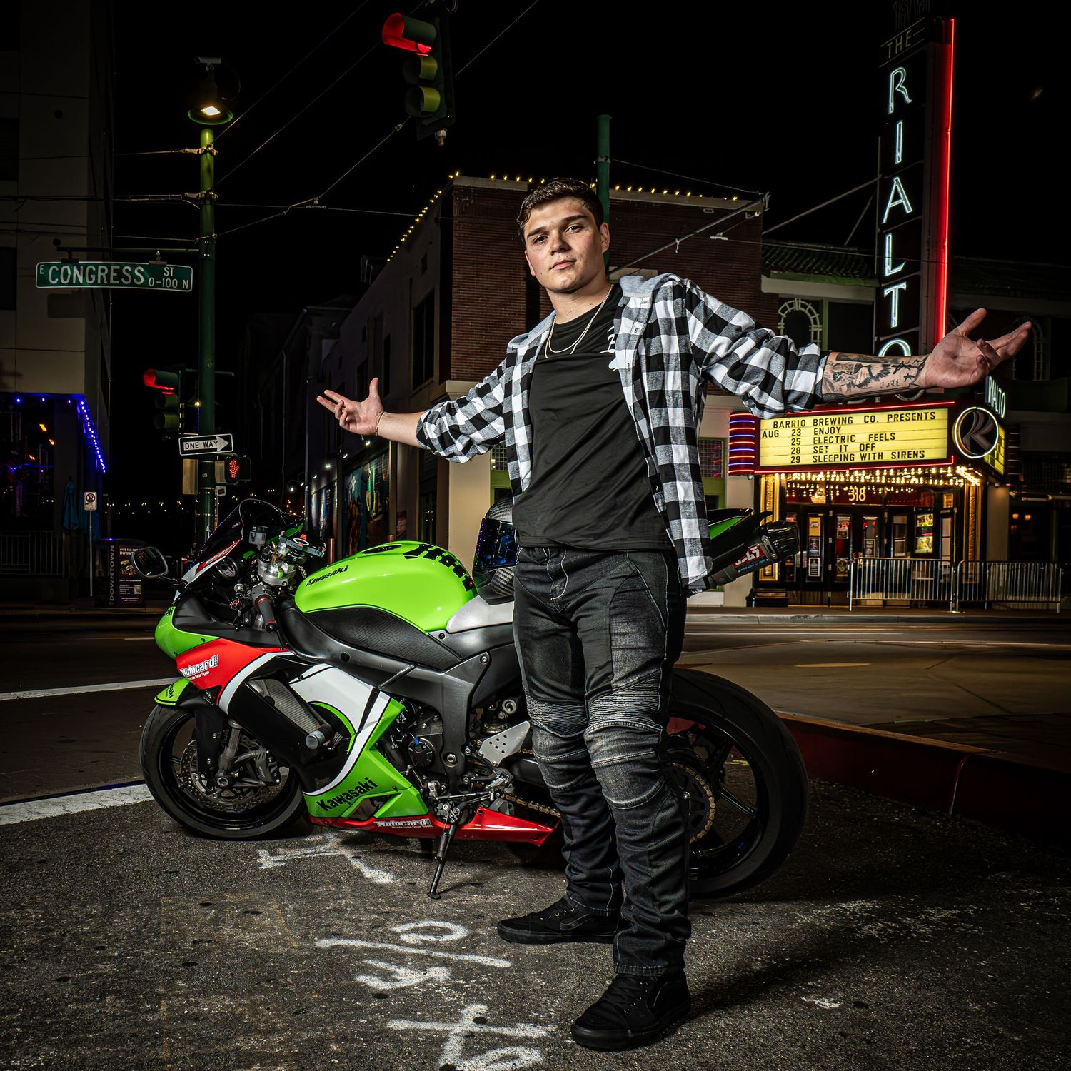 Young man in casual attire posing with a green sport motorcycle on a nighttime city street.