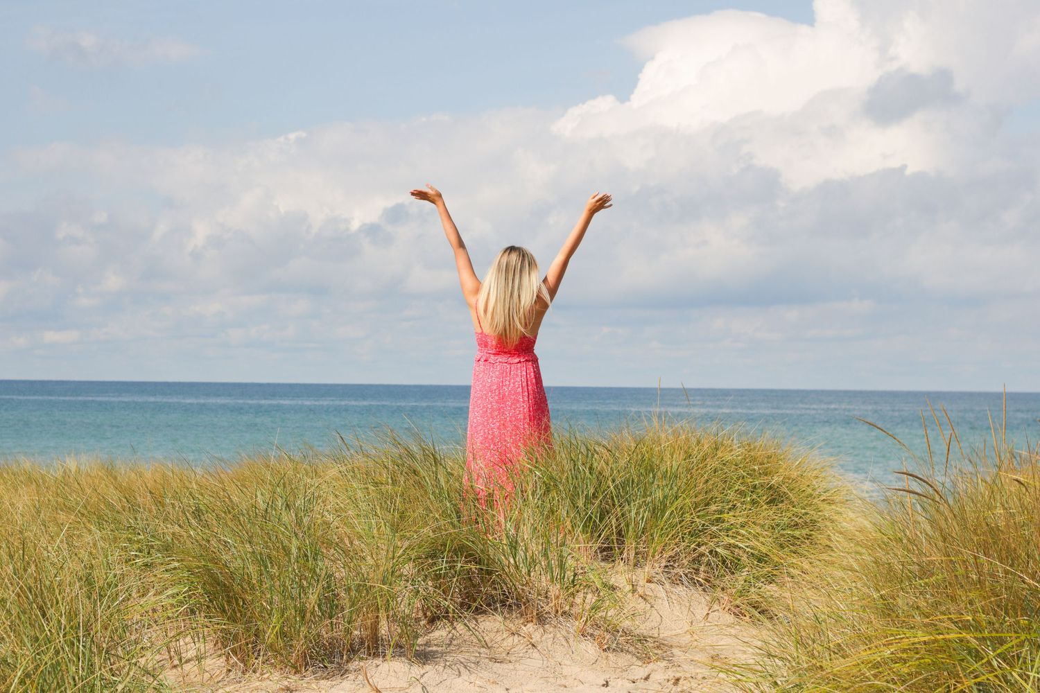 Figure in pink dress stands triumphantly on beach dunes against ocean backdrop.