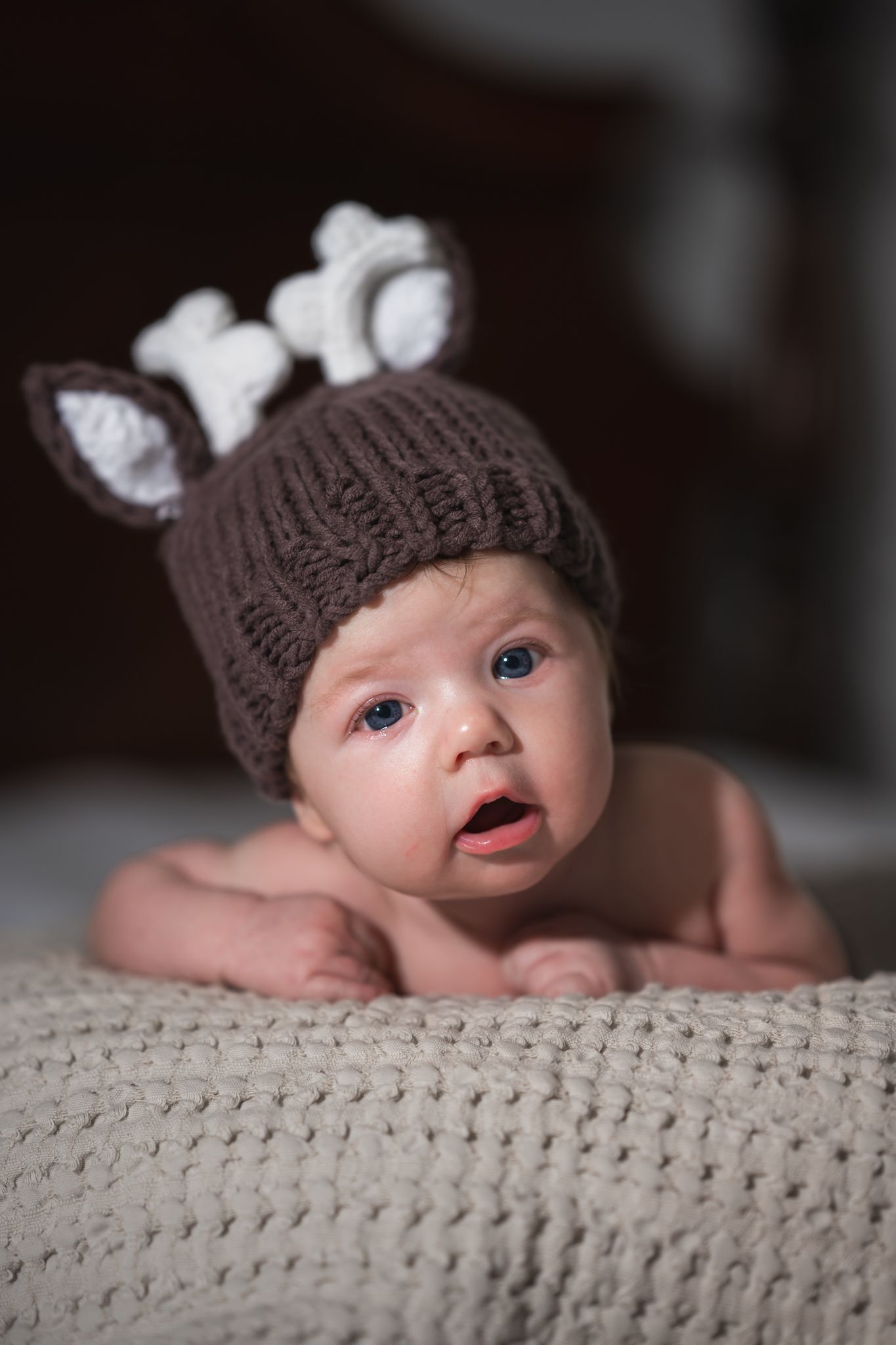 Baby wearing knit deer hat laying on textured grey blanket.