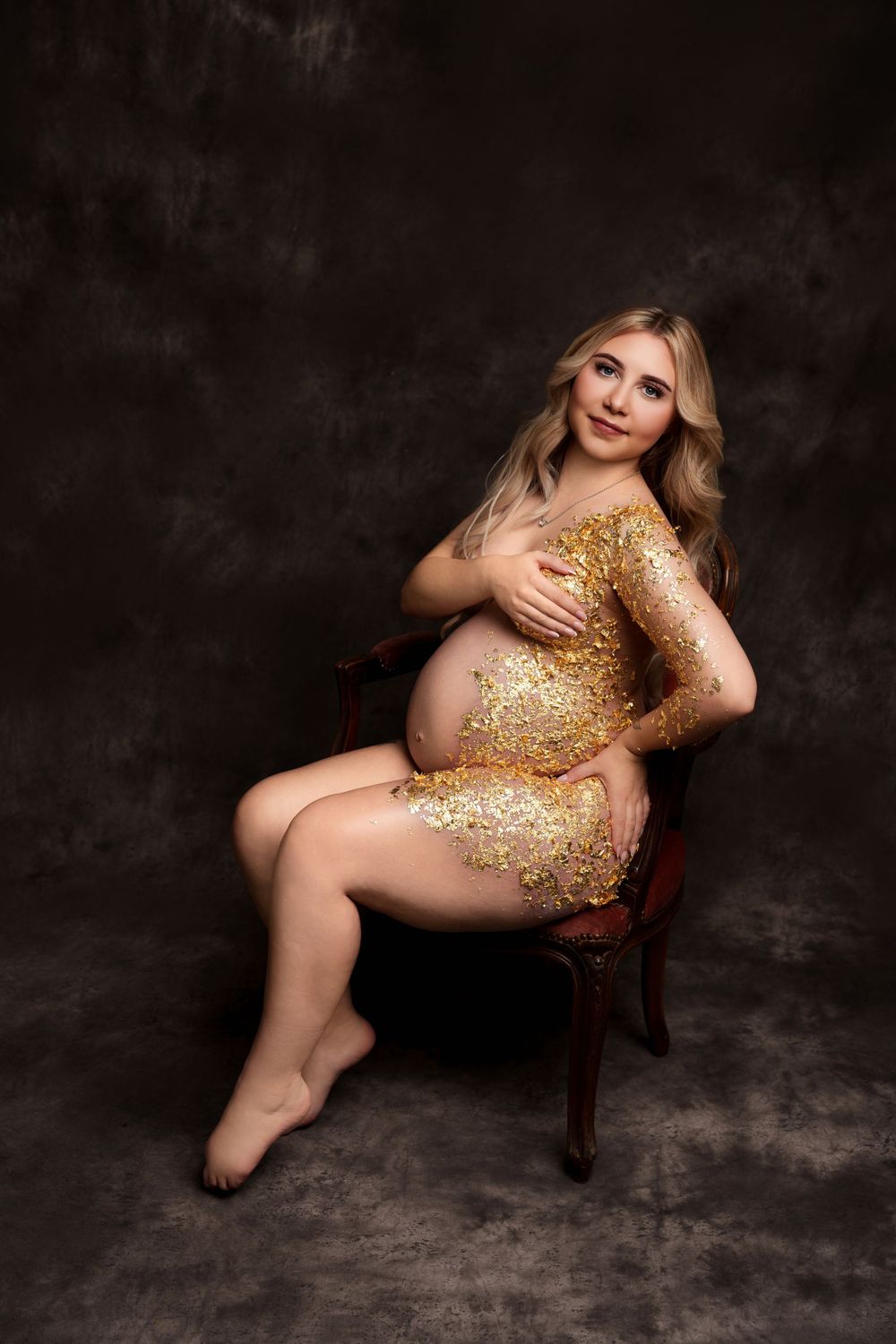 Glamorous maternity portrait with gold glitter dress in antique chair setting.