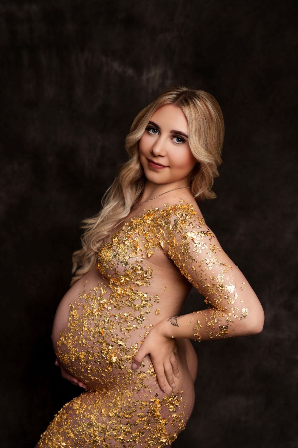 Elegant maternity photo with sparkly gold body art against dark backdrop.