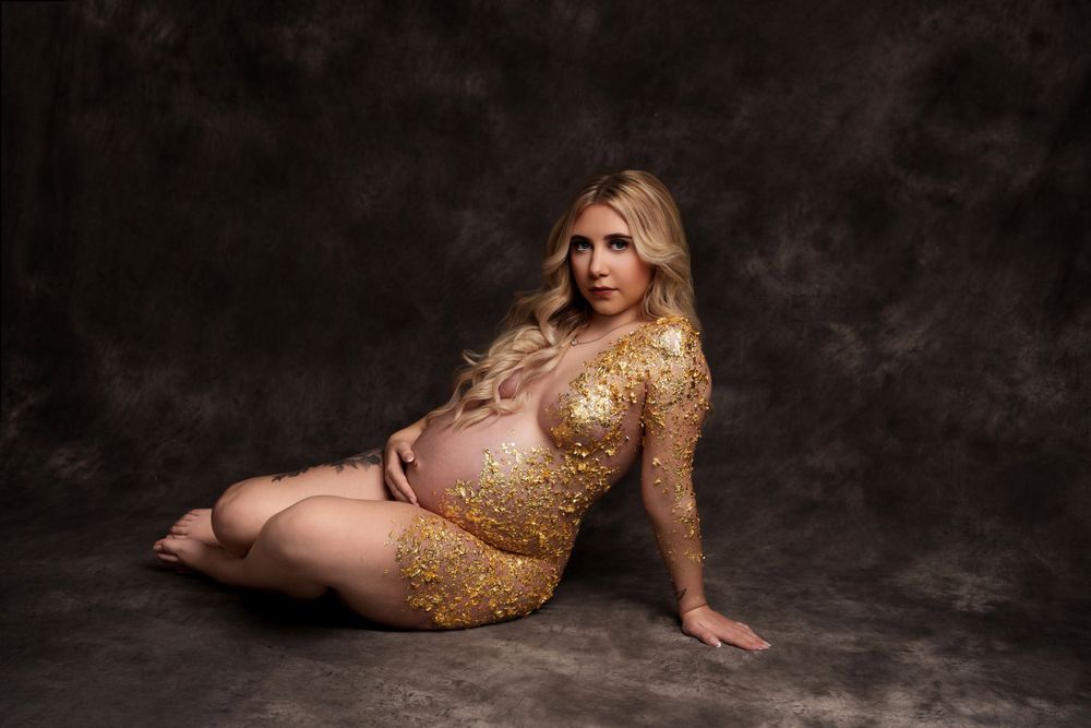 Maternity portrait with gold glitter bodysuit against dark background.
