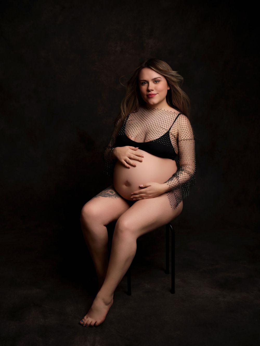 Maternity boudoir photo with sheer mesh top on dark background.
