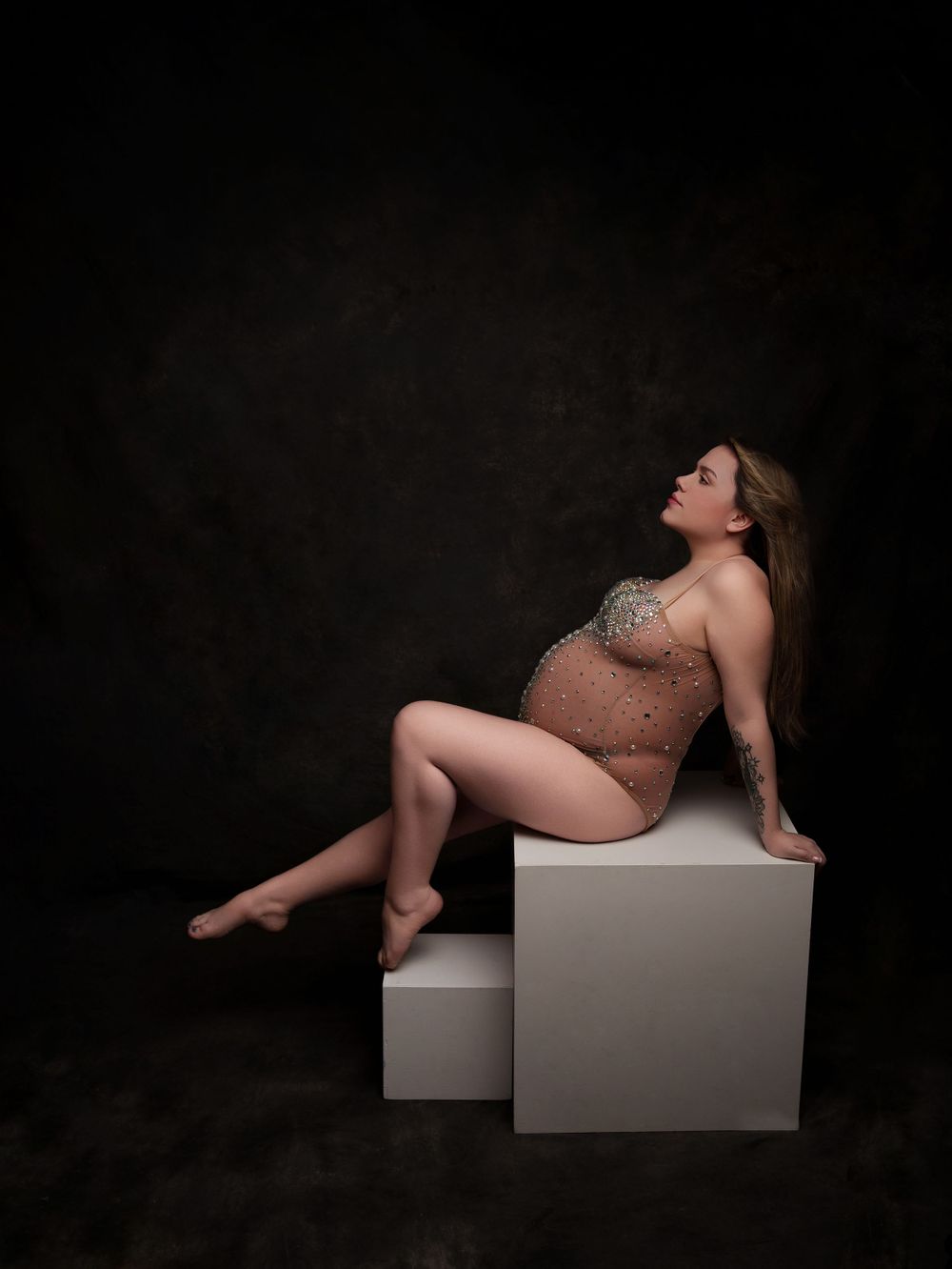 Artistic maternity pose on white geometric blocks against dark backdrop.
