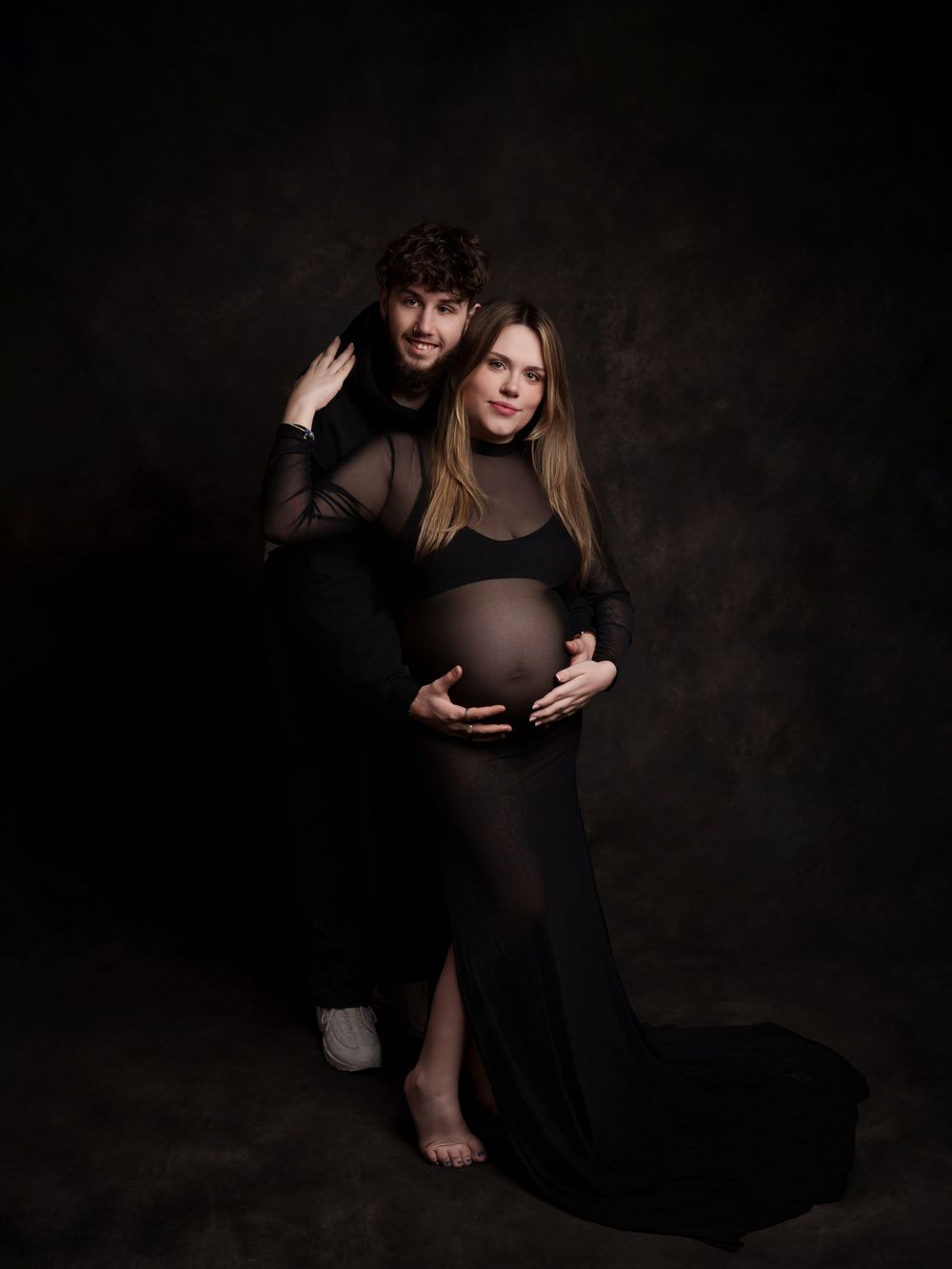 Artistic maternity portrait in dark tones featuring couple in black clothing against moody background.