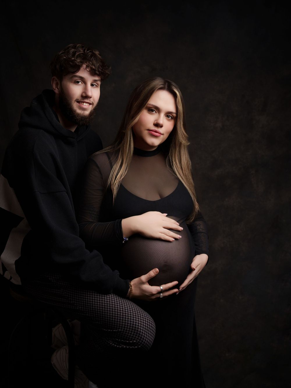 Intimate maternity portrait of couple in dark clothing against moody background.