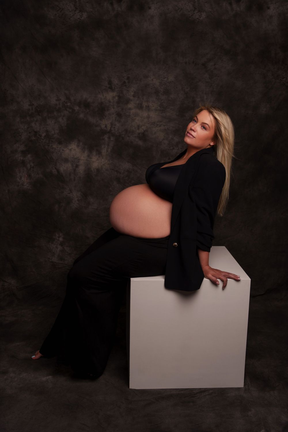 Maternity photo featuring woman in black outfit posing against white cube on dark background.