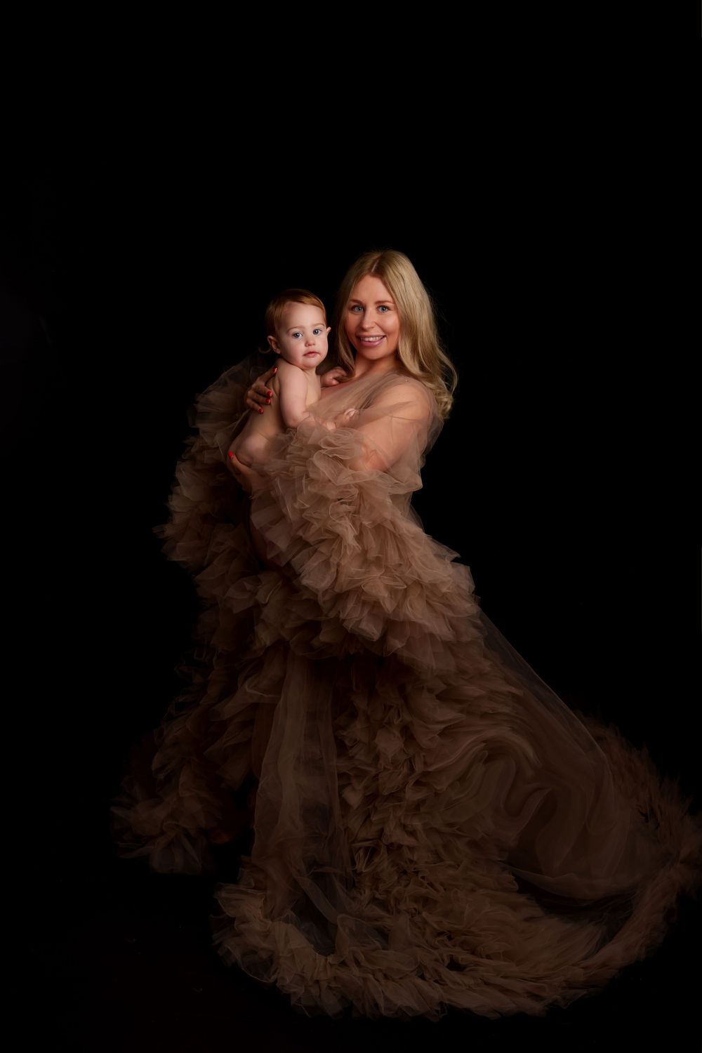 Mother and baby portrait in flowing brown tulle gown against black background.