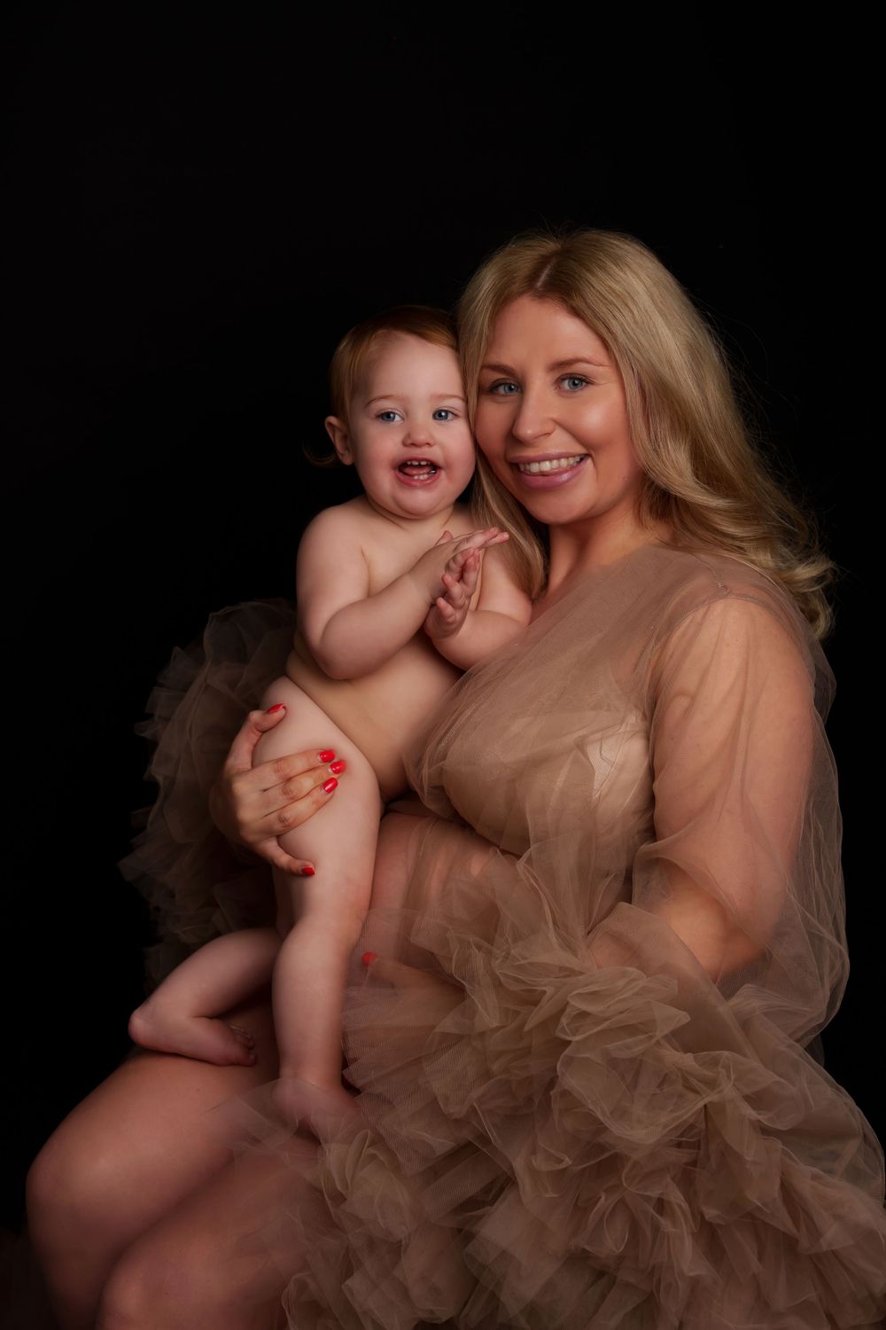 Mother and baby in ethereal tulle dress pose against dark background for intimate portrait session.