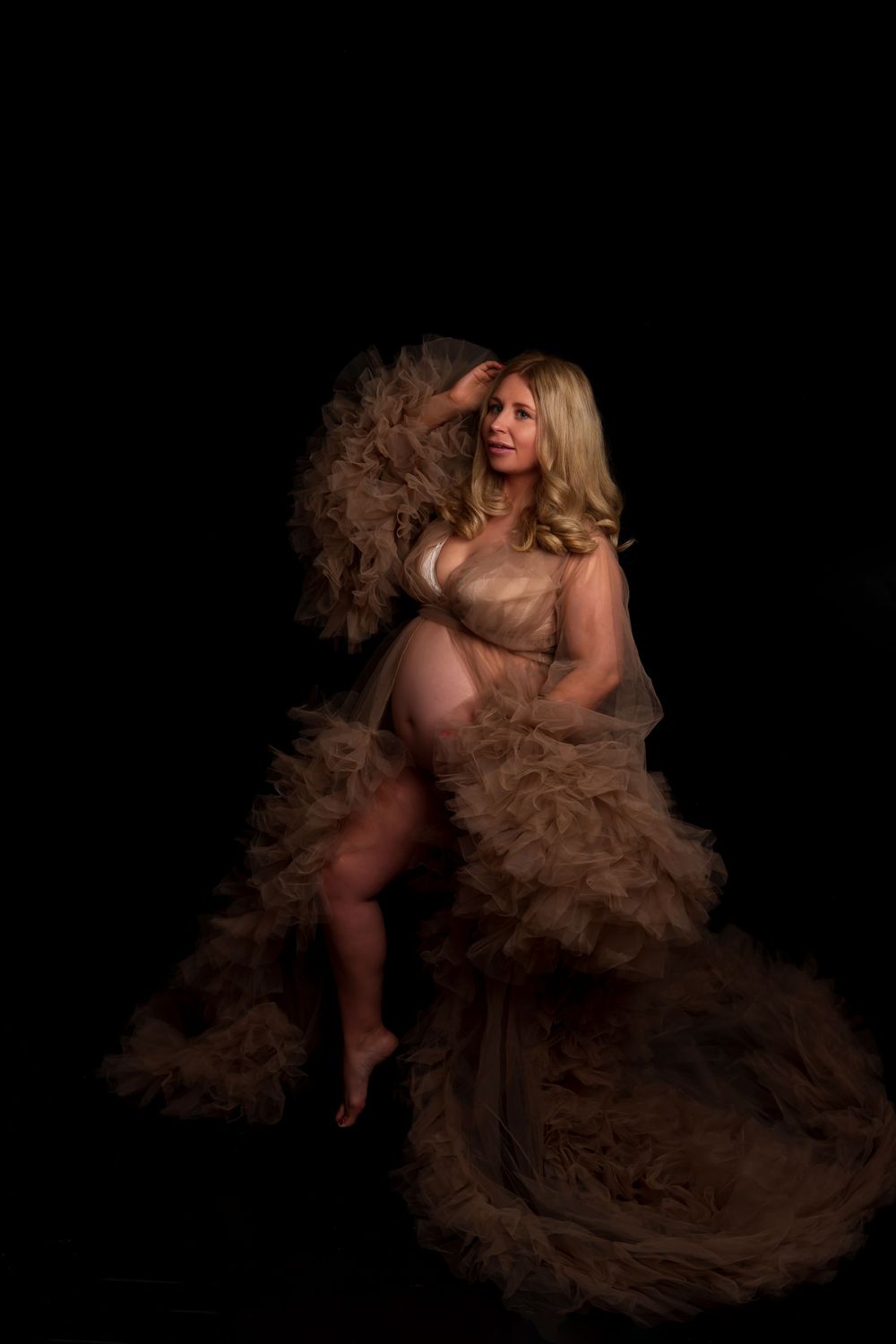 Maternity portrait featuring flowing champagne tulle gown against black background.