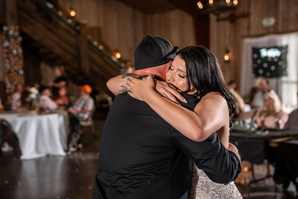 A couple shares an intimate dance moment at a wedding reception in a rustic venue with twinkling lights.