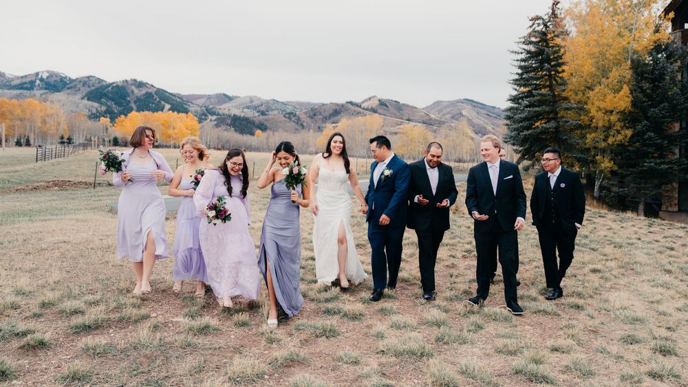 Wedding party in formal attire walks together across scenic mountain meadow with autumn colors at sunset.