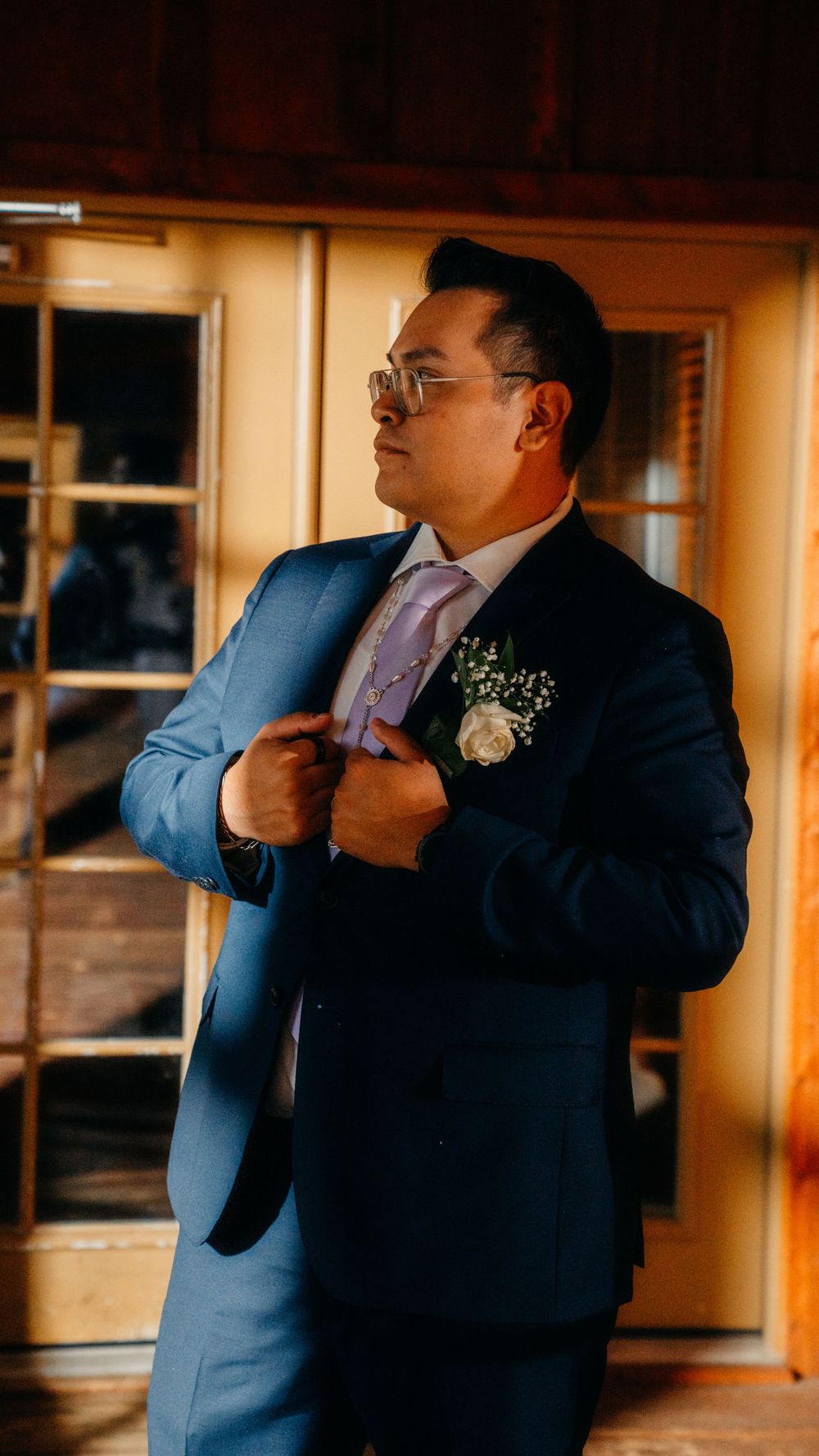A person in a navy blue suit adjusts their tie while getting ready in a warm-lit room.