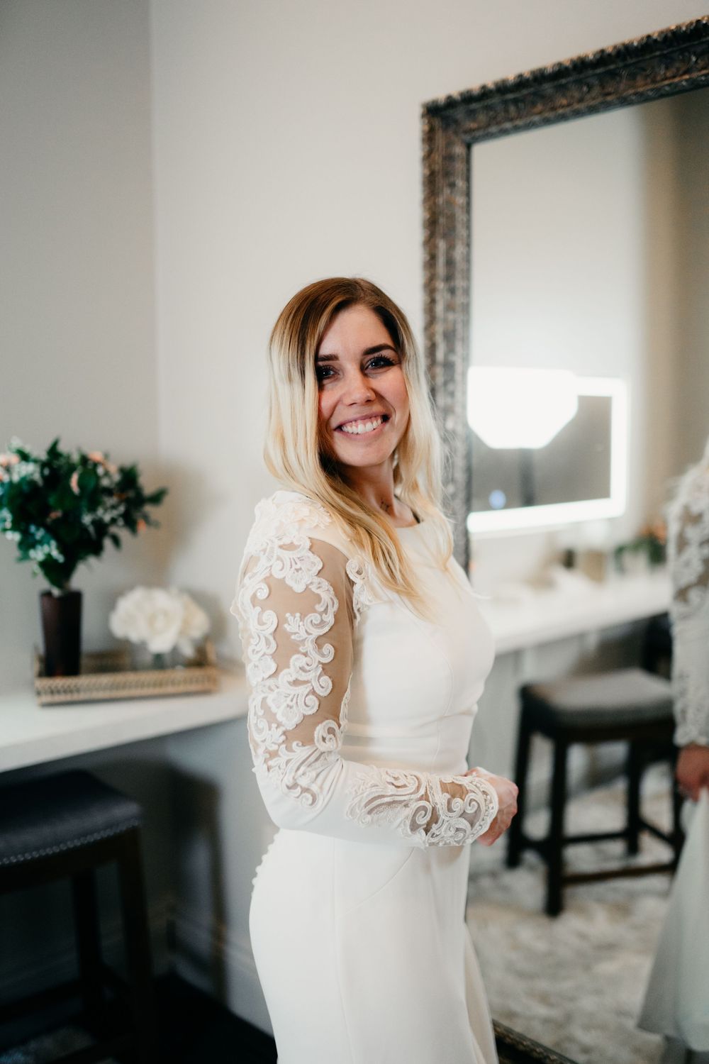 A bride in a long-sleeved lace wedding gown poses in a bright white room with elegant decor.