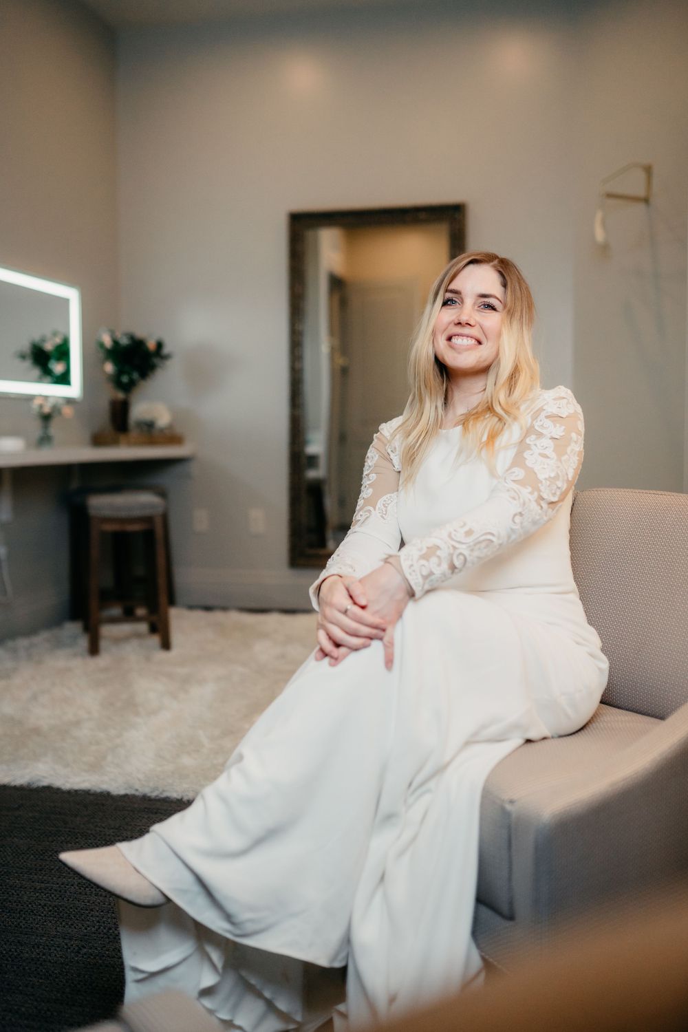 A bride in a flowing white lace gown sits casually on a couch in an indoor setting.