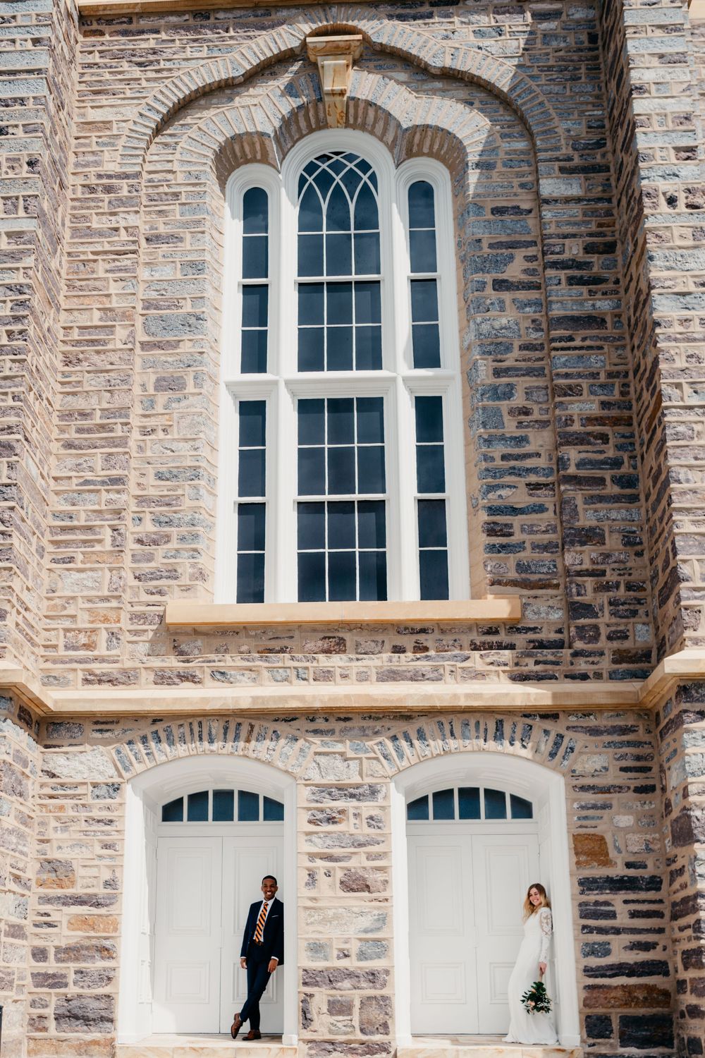 Stone church facade with arched windows and multiple figures standing in white doorway openings.