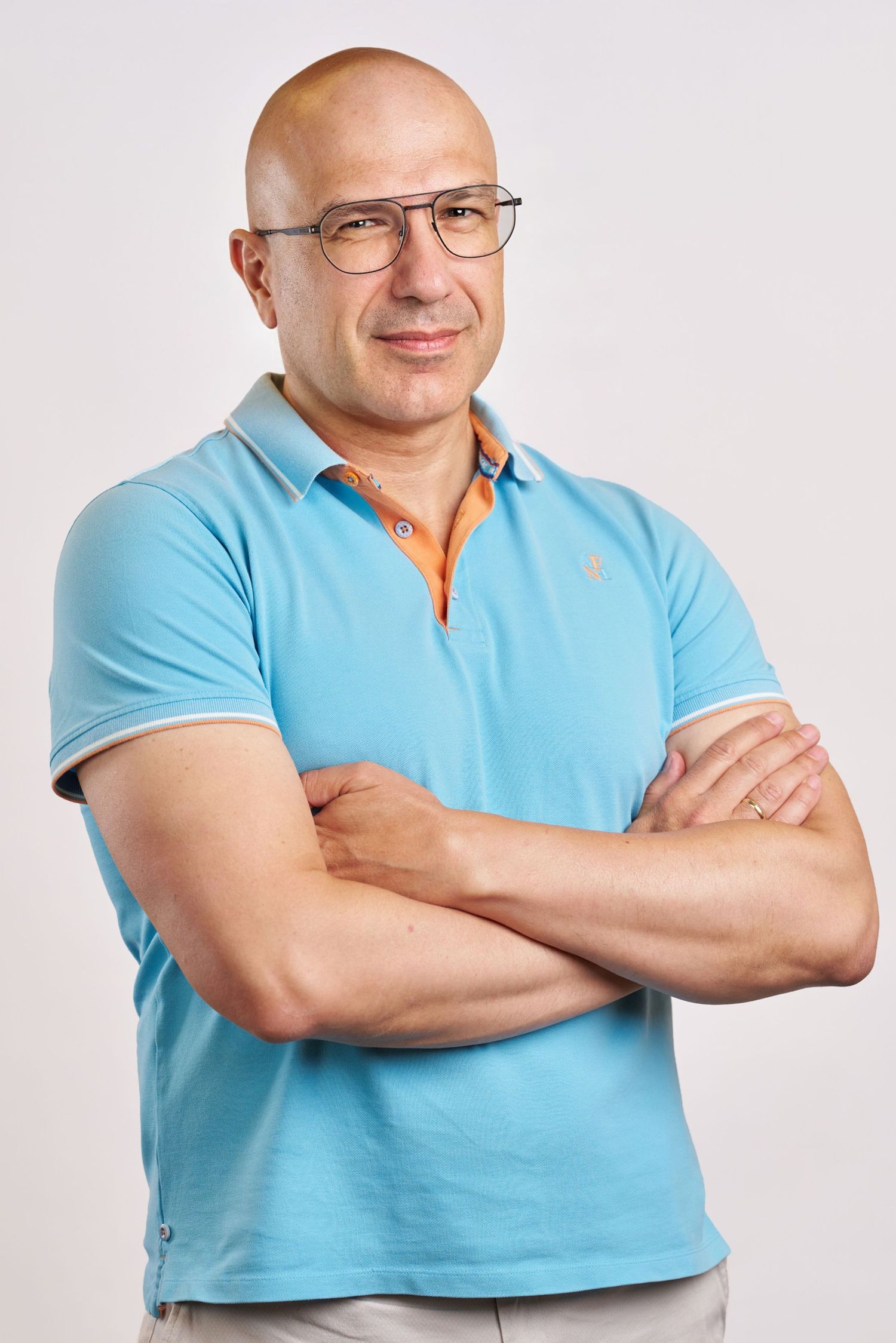 Bald middle-aged man with glasses wearing a light blue polo shirt, standing with arms crossed against a white background.