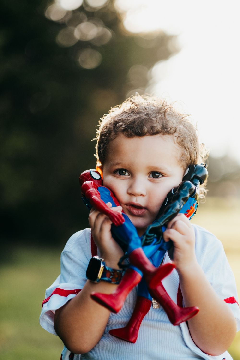 A young boy with curly hair holding his favorite toys, a Spider-Man and a Venom up to his cheeks.