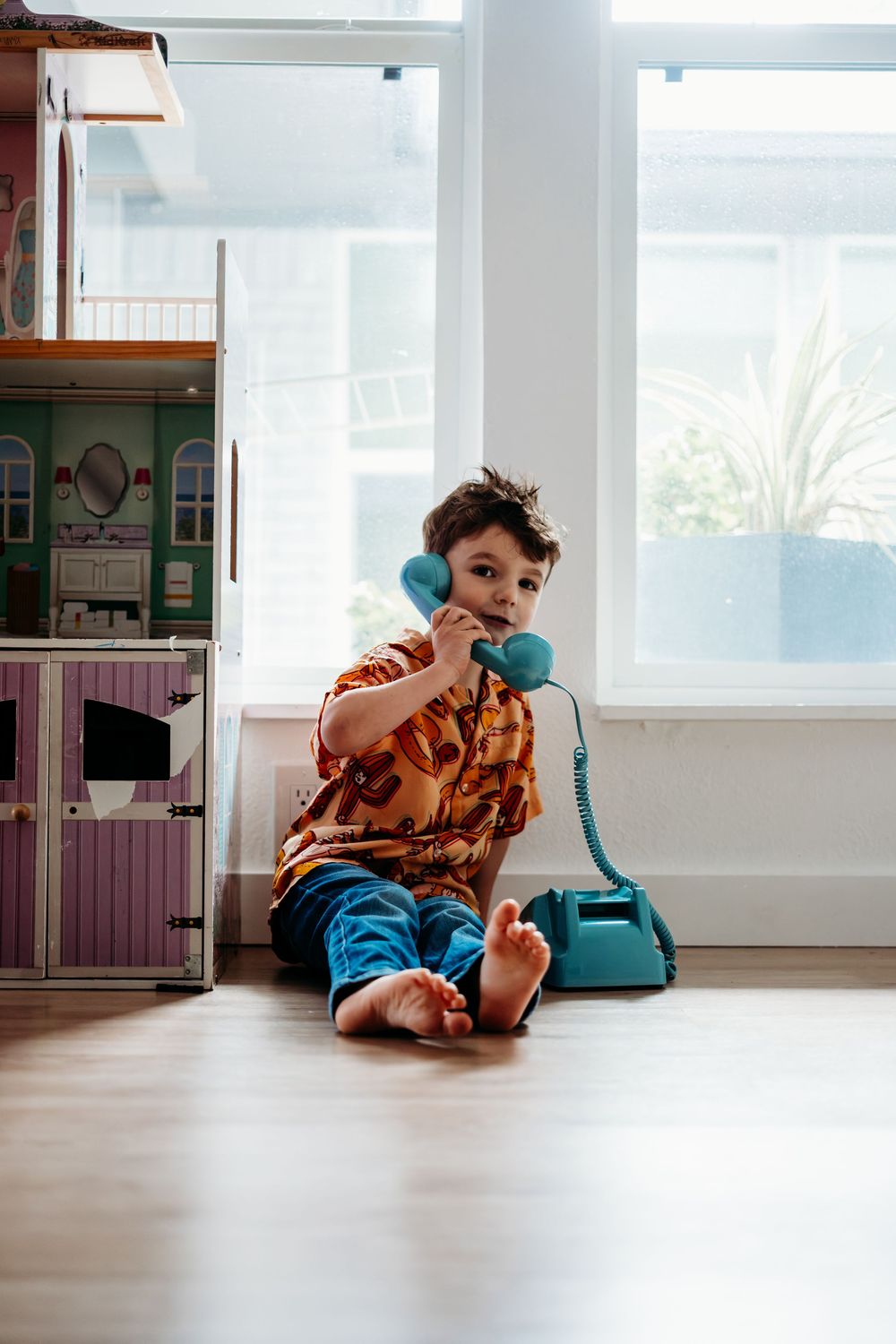 A young boy is pretending to talk on an old school corded blue telephone while sitting on the floor in front of a bank of windows.