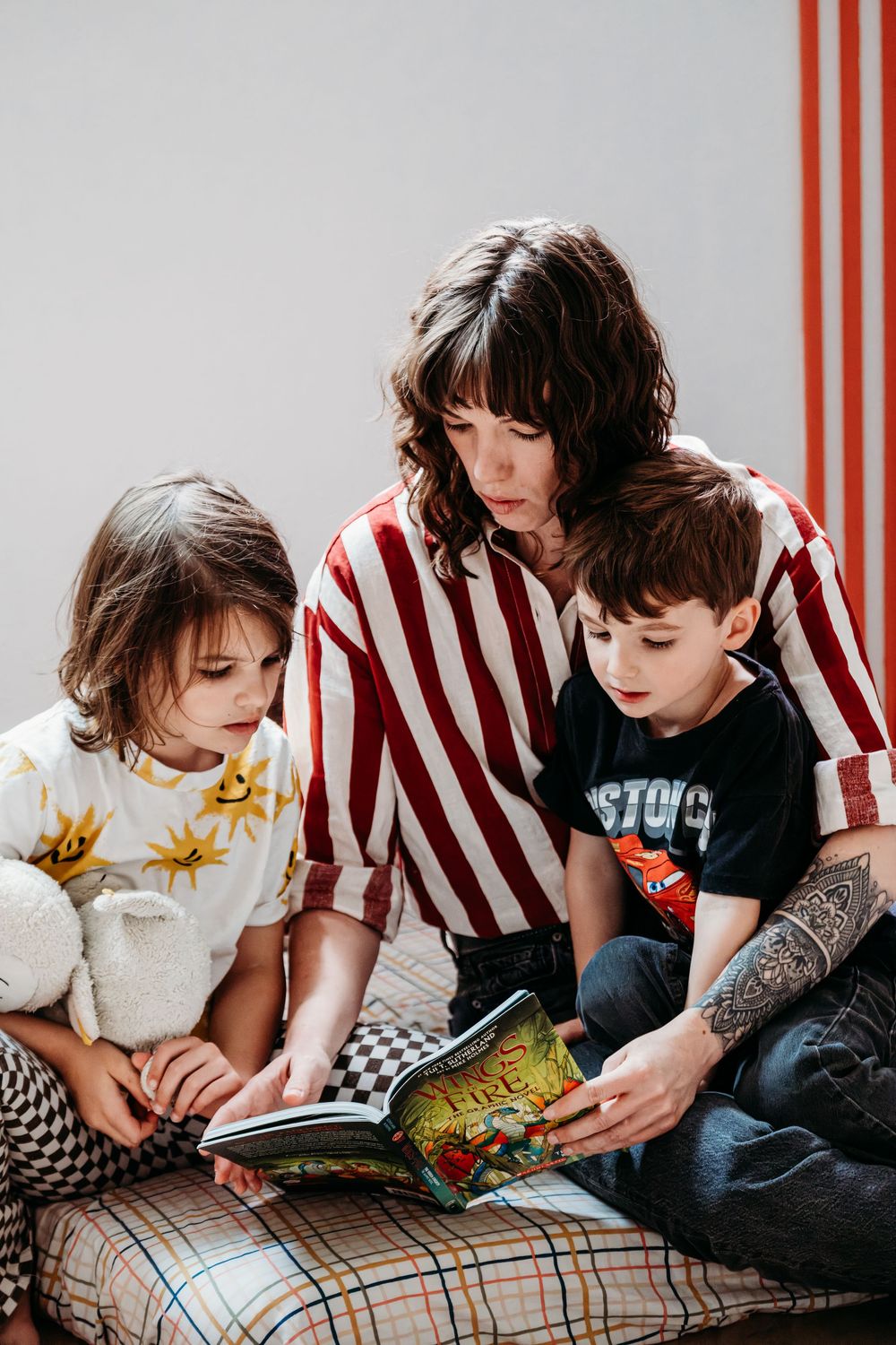 A mother and her 2 young children are reading a book together in a brightly lit room