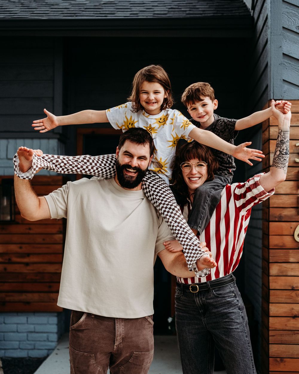 A daughter sits on her dad's shoulders, and a son sits on his moms shoulders in front of their home with their arms open wide and big smiles