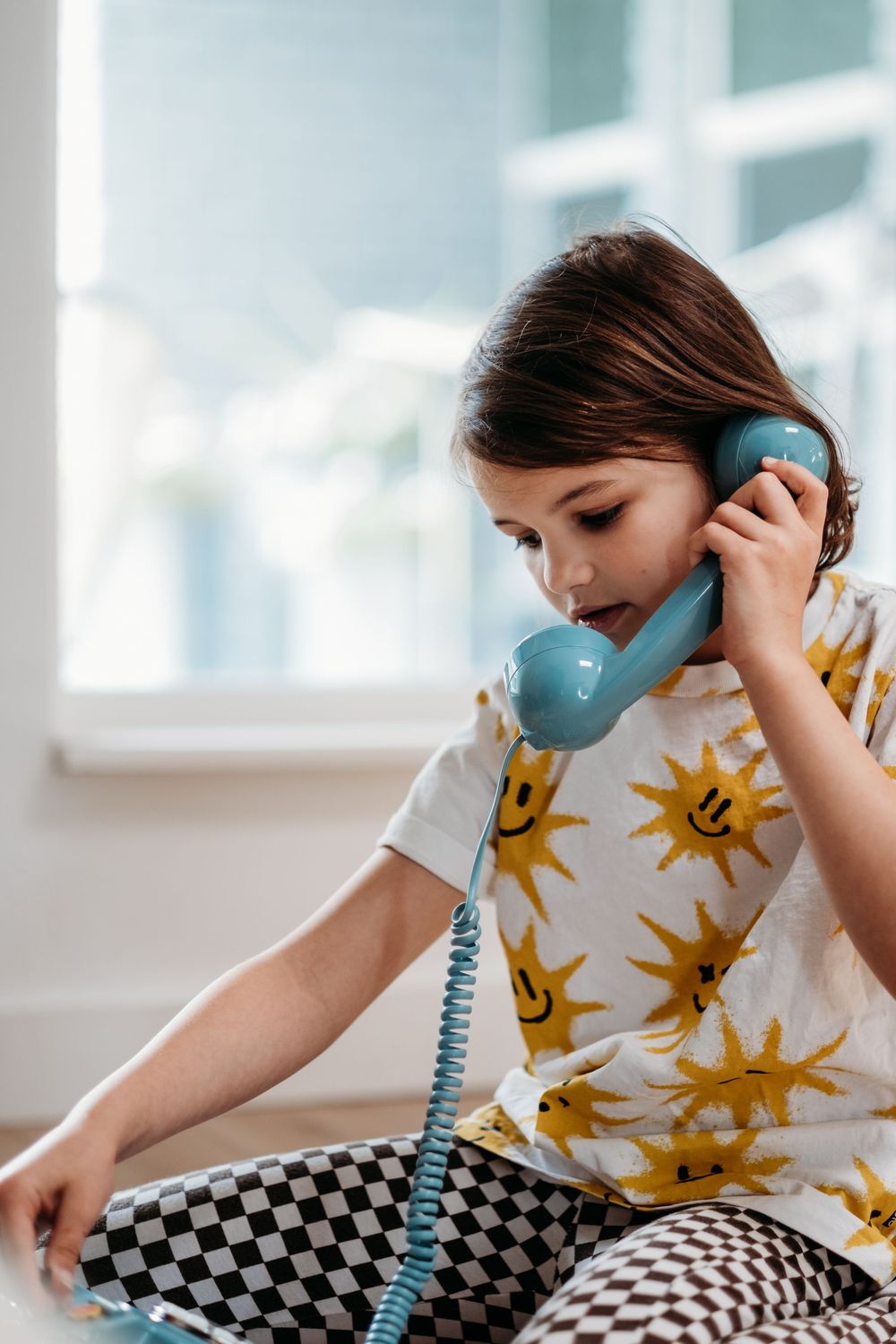 A young girl wearing a shirt with yellow suns is holding an old school corded blue phone to her ear and pretending to be on a phone call