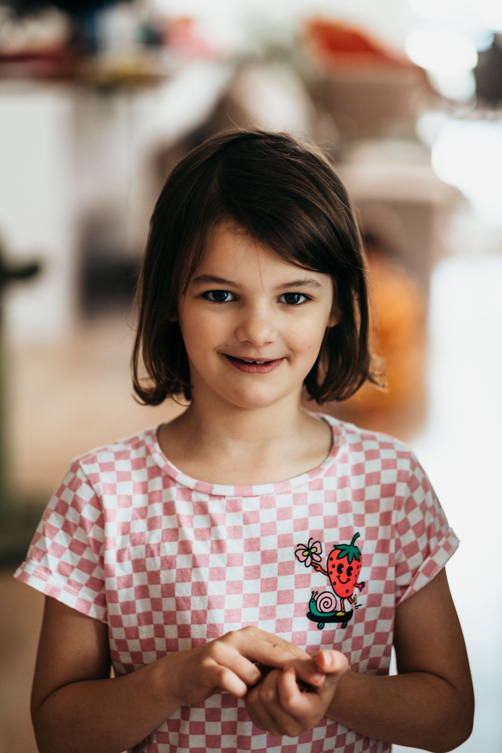 A young girl with short brown hair and a pink and white shirt is smiling at the camera while holding a small trinket in her hand.