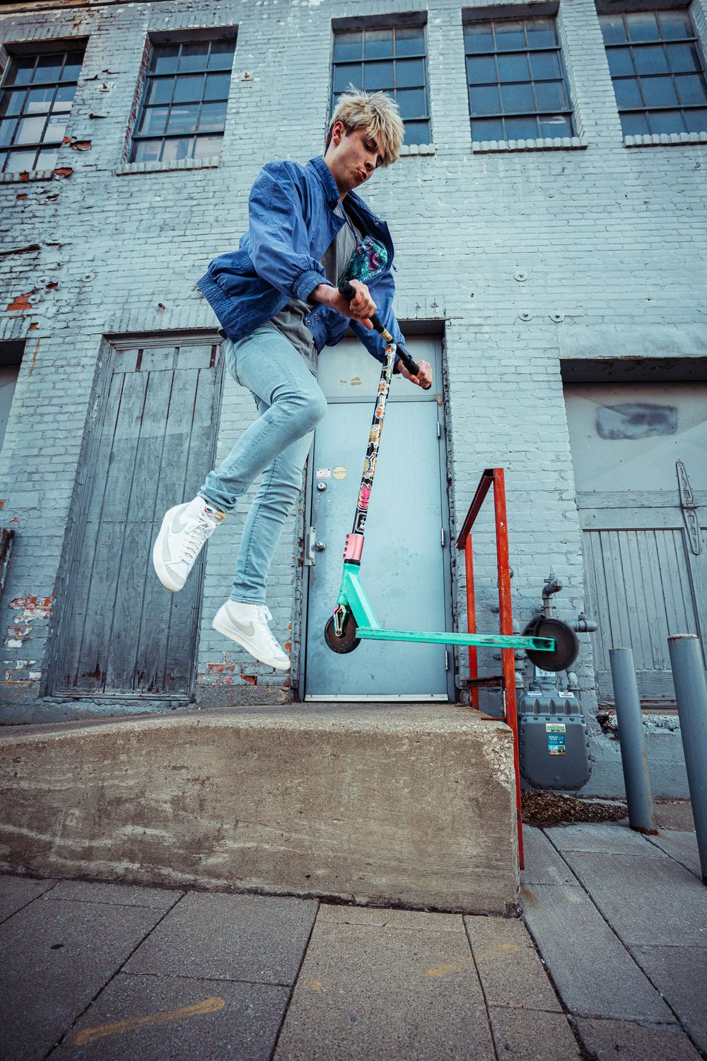 Action shot of person jumping with teal scooter in front of industrial building.